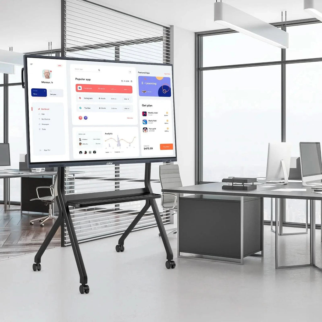 JAV Heavy Duty Rolling TV Stand in a modern office. This black mobile stand features lockable wheels, a shelf, and is shown supporting a large display screen with an application dashboard. Ideal for office presentations, meetings, and interactive workspaces.