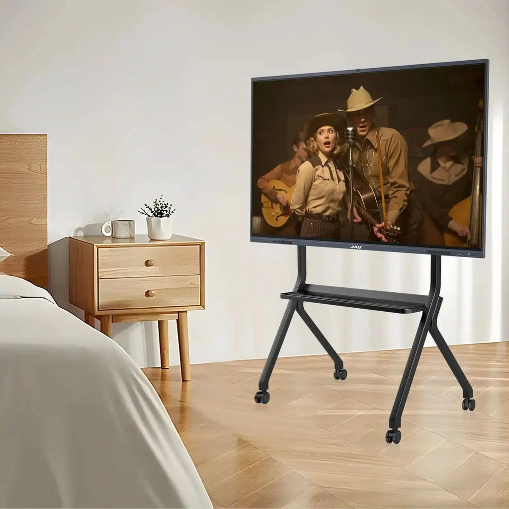 JAV Mobile Rolling TV Stand in a bedroom. This black portable stand with wheels and shelf is shown in a home bedroom setting, displaying a musical performance on screen. Ideal for bedroom TVs and flexible home entertainment setups.