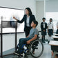 A wheelchair-using student operates a lowered smart board on an electric lift stand, guided by a teacher, demonstrating accessibility for diverse needs.