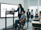 A wheelchair-using student operates a lowered smart board on an electric lift stand, guided by a teacher, demonstrating accessibility for diverse needs.