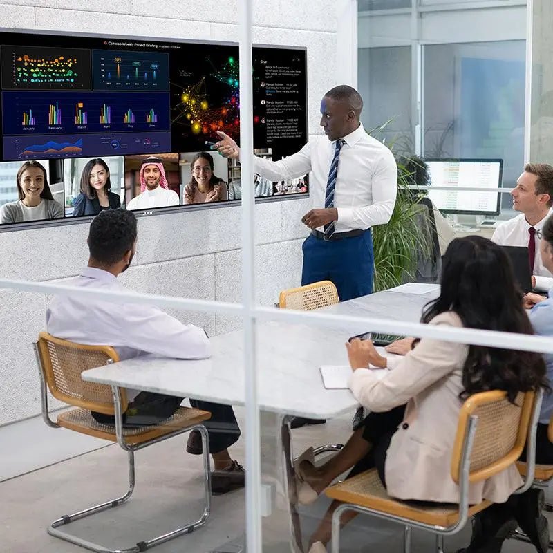 Team using a JAV H15 interactive display for a presentation in a hybrid meeting, with one person standing and pointing at data and video participants on screen.