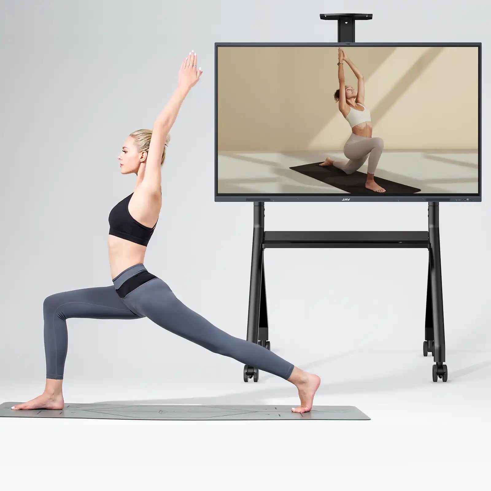 Woman performing yoga pose while following instructional video on a large JAV smart board positioned on a mobile stand.