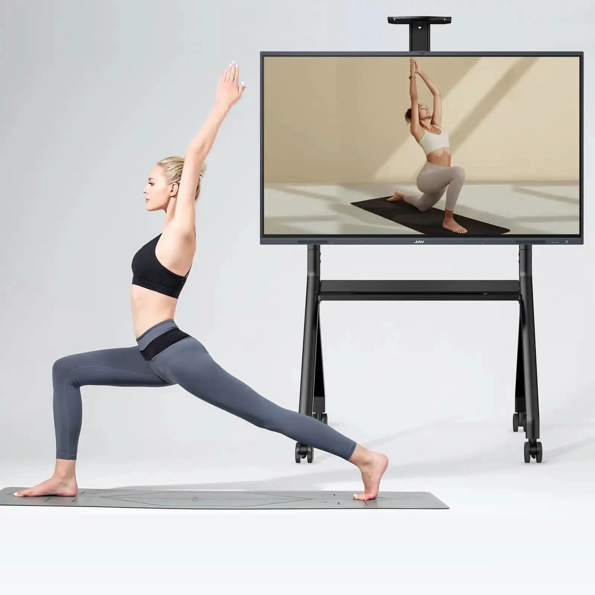 Woman performing yoga pose while following instructional video on a large JAV smart board positioned on a mobile stand.